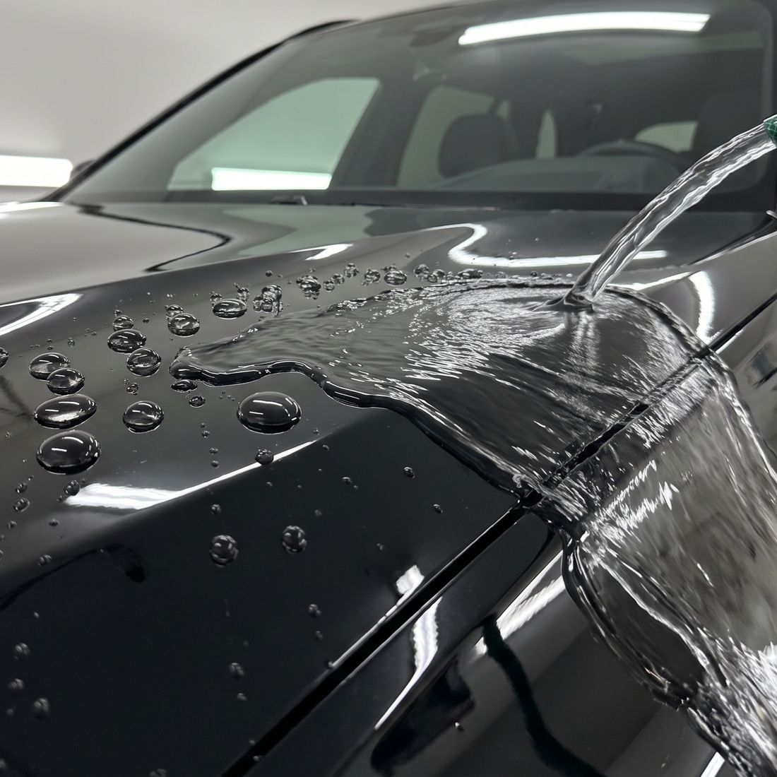 Water from a hose running off the glossy black hood of an SUV in a white garage, round beads standing on the paint beside it.