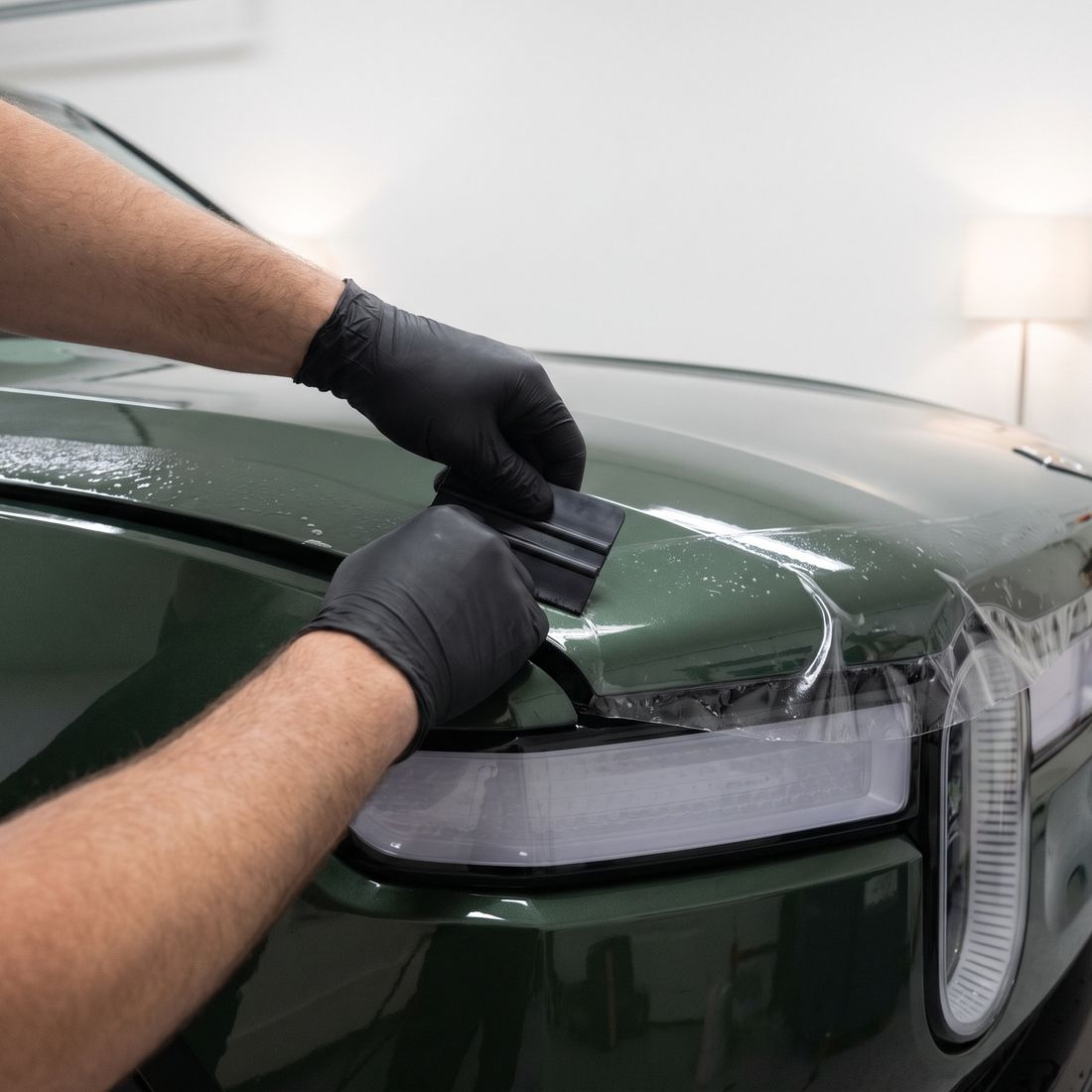 Gloved hands pressing a squeegee over clear film on the front of a dark green SUV's hood, the film edge tucked beside the headlight.