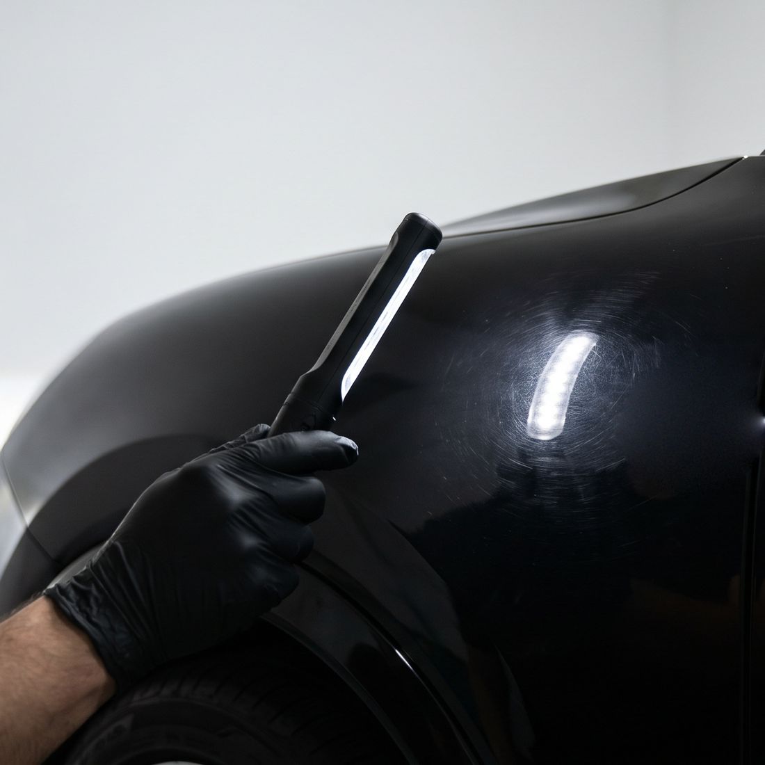 A gloved hand holding a slim LED inspection light against the black fender of a car, the light picking out fine circular scratches in the paint.