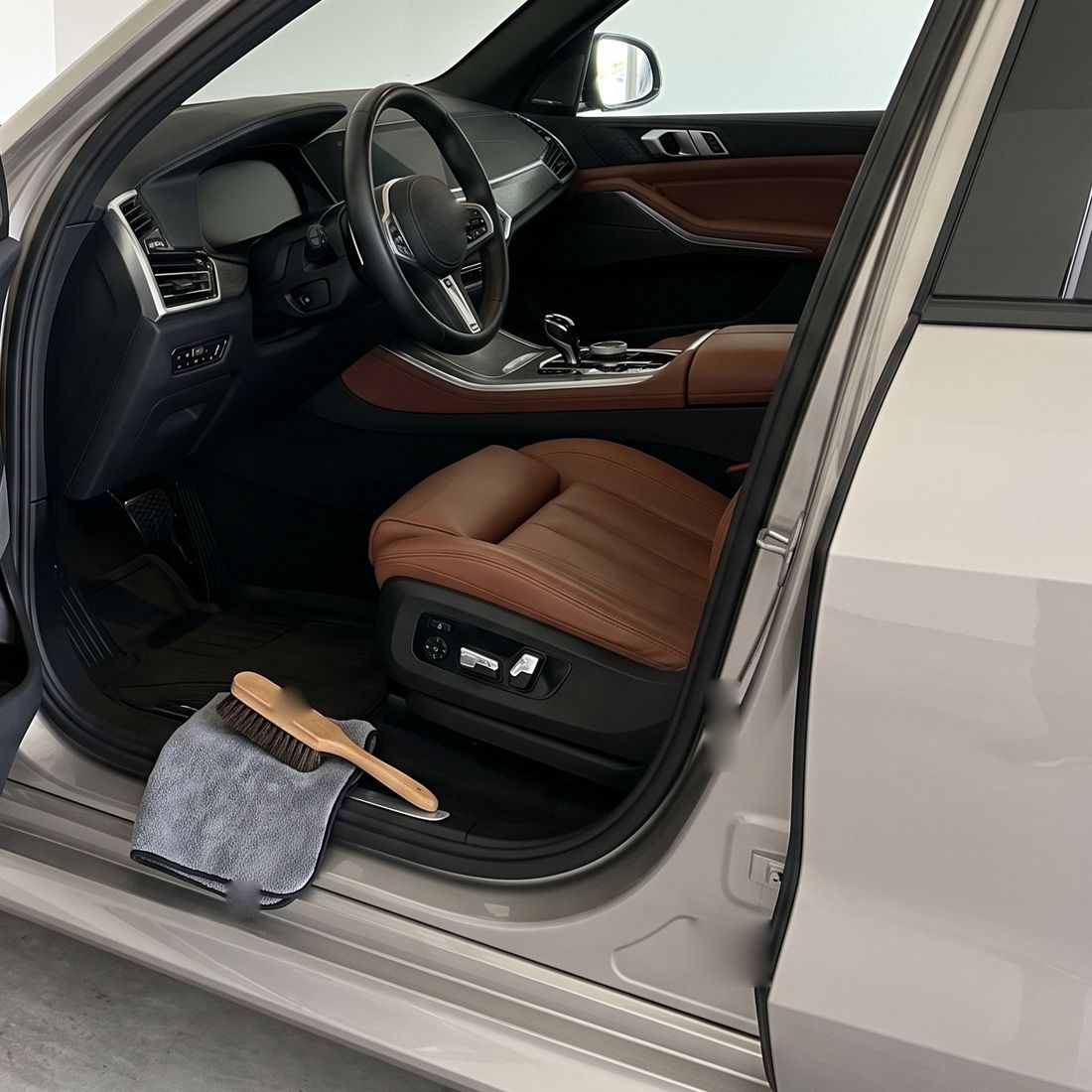 Cognac leather front seats seen through the open driver's door of an SUV in a white garage, a gray towel and a horsehair brush on the door sill.