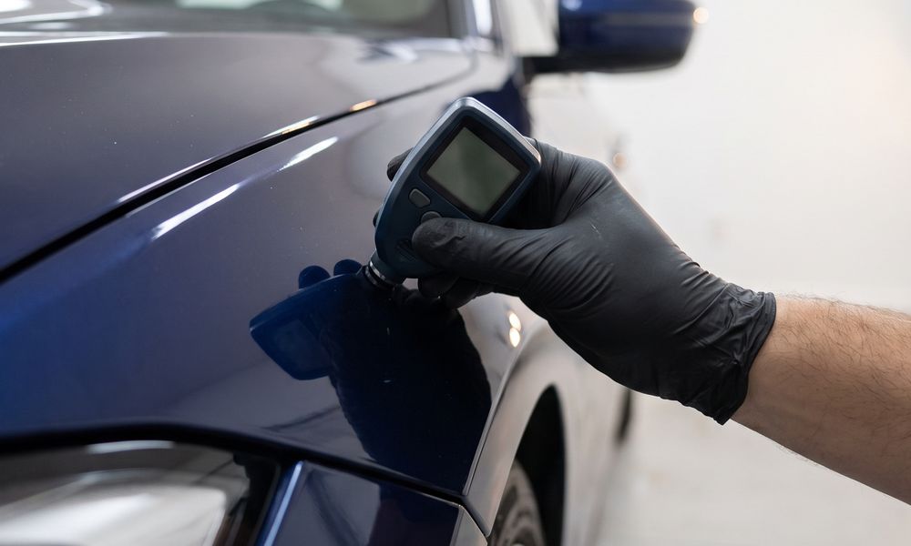 A gloved hand holding a paint thickness gauge against the front fender of a dark blue car in a white garage.