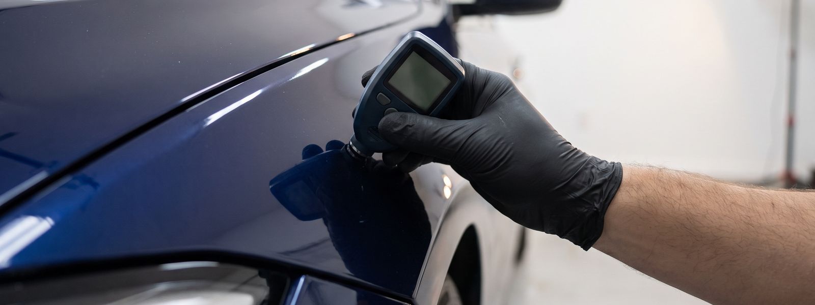 A gloved hand holding a paint thickness gauge against the front fender of a dark blue car in a white garage.