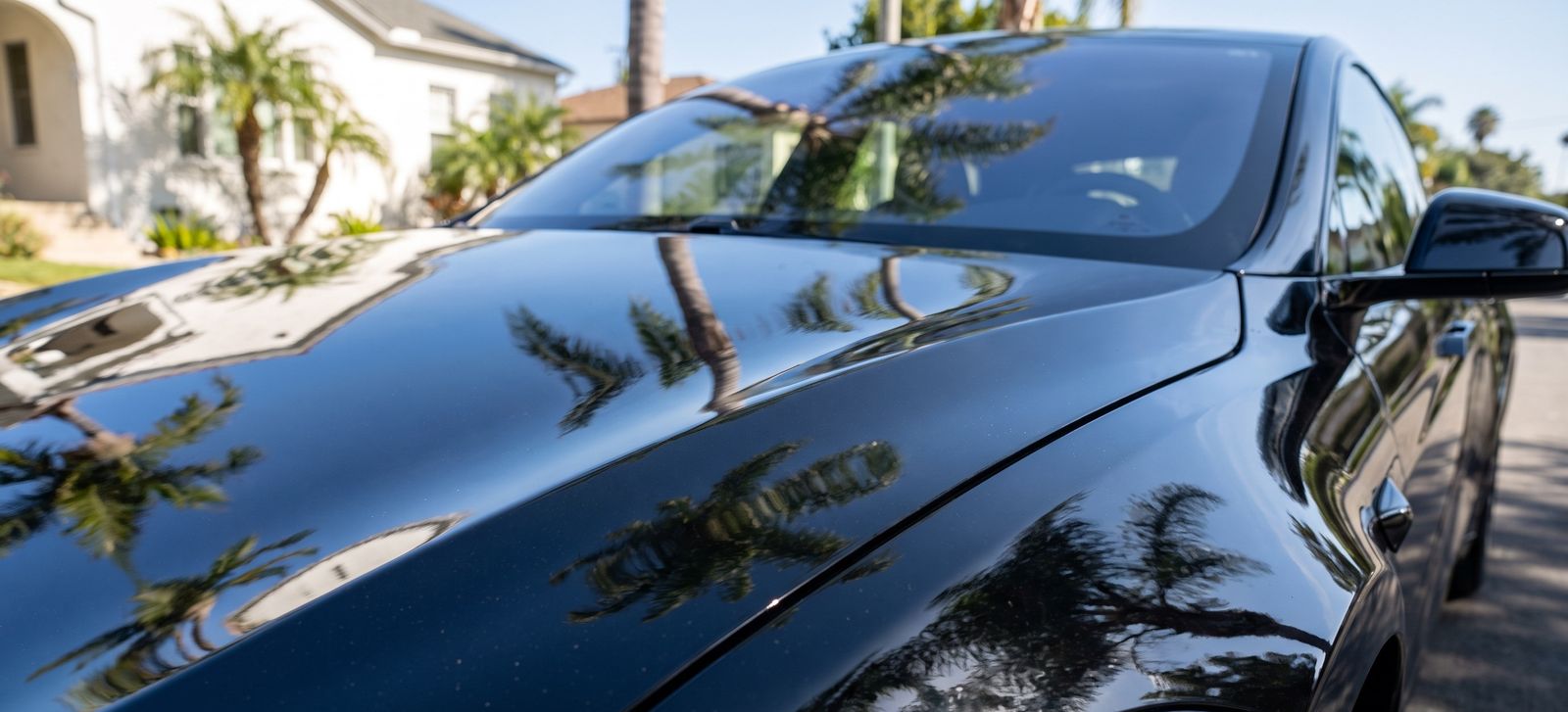The polished black hood of a sedan parked beside a white stucco house, mirroring palm fronds and a clear blue sky.