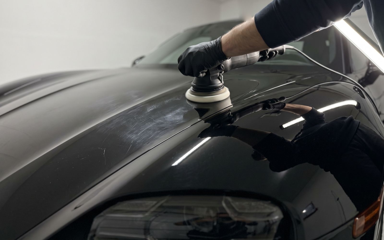 A gloved hand running a polisher over the hood of a black car in a white garage, fine swirls on one side of the pad and clear, polished paint on the other.